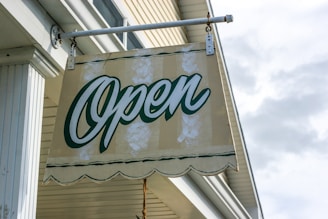 A cream-colored sign with the word 'Open' written in green script hangs outside a building. The sign has a scalloped bottom edge and is attached to a bracket protruding from the building. The background shows part of the building's exterior, with white siding and architectural details, and a slightly cloudy sky.