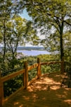 A wooden deck bordered by a railing overlooks a scenic view. Lush green trees frame the scene, with a distant river and landscape visible through the foliage. Sunlight creates playful patterns of light and shadow on the deck surface.