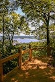 A wooden deck bordered by a railing overlooks a scenic view. Lush green trees frame the scene, with a distant river and landscape visible through the foliage. Sunlight creates playful patterns of light and shadow on the deck surface.