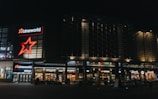 A modern commercial complex at night featuring illuminated signs for various establishments including a cinema and multiple restaurants. The architecture includes glass and metal elements with bright neon signage.