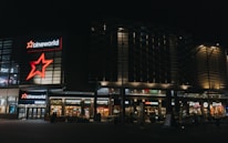 A modern commercial complex at night featuring illuminated signs for various establishments including a cinema and multiple restaurants. The architecture includes glass and metal elements with bright neon signage.