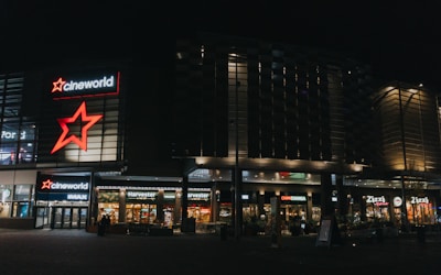 A modern commercial complex at night featuring illuminated signs for various establishments including a cinema and multiple restaurants. The architecture includes glass and metal elements with bright neon signage.