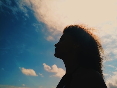A silhouette of a person with curly hair against a bright sky. The clouds are sparse, and the sun is shining behind, creating a halo effect around the head.