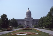 A large, domed government building centered in the image, surrounded by well-maintained gardens and trees. In the foreground, a path lined with colorful flower beds leads up to the building. A yellow school bus is parked on the left side of the road, under the shade of the trees.