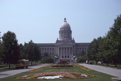 A large, domed government building centered in the image, surrounded by well-maintained gardens and trees. In the foreground, a path lined with colorful flower beds leads up to the building. A yellow school bus is parked on the left side of the road, under the shade of the trees.