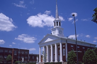 A red-brick building with a white facade and a tall steeple topped with a cross. The facade features large columns and clock faces on either side. The sky is bright blue with scattered clouds, and there are trees and streetlights around the building.