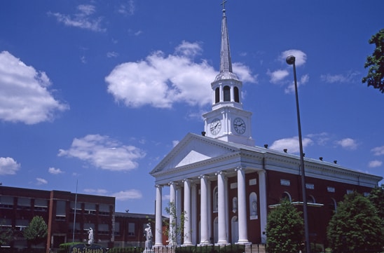 A red-brick building with a white facade and a tall steeple topped with a cross. The facade features large columns and clock faces on either side. The sky is bright blue with scattered clouds, and there are trees and streetlights around the building.