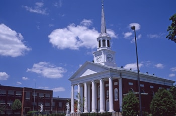A red-brick building with a white facade and a tall steeple topped with a cross. The facade features large columns and clock faces on either side. The sky is bright blue with scattered clouds, and there are trees and streetlights around the building.