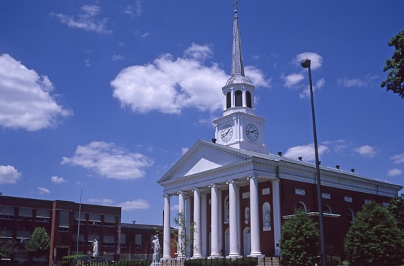 A red-brick building with a white facade and a tall steeple topped with a cross. The facade features large columns and clock faces on either side. The sky is bright blue with scattered clouds, and there are trees and streetlights around the building.