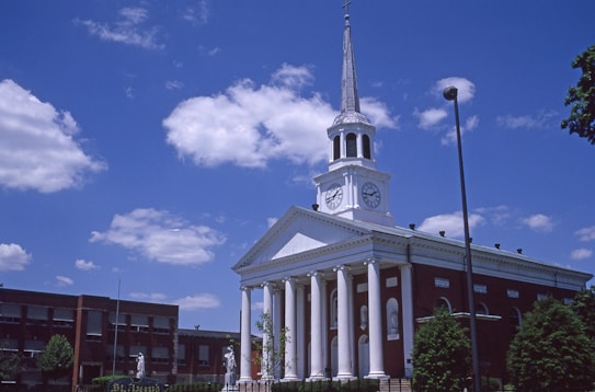 A red-brick building with a white facade and a tall steeple topped with a cross. The facade features large columns and clock faces on either side. The sky is bright blue with scattered clouds, and there are trees and streetlights around the building.