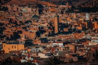 A dense cityscape with numerous beige and orange buildings, many topped with satellite dishes. The rooftops are a mixture of flat and sloped designs. A tall mosque minaret stands out in the center among the buildings. The scene is set against a backdrop of rolling hills and sparse vegetation, creating a warm, rustic atmosphere.