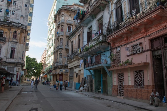 A vibrant street scene featuring colorful, weathered buildings with balconies adorned with plants and laundry. People are casually walking along the street, which is lined with a variety of architectural styles. The atmosphere feels lively and authentic, with a bright sky providing contrasting light and shadows.
