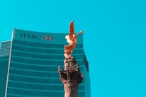 A golden angel statue stands prominently on top of a tall column against a backdrop of a modern glass skyscraper with the HSBC logo visible. The sky is clear and bright blue, providing a striking contrast to the statue and building.