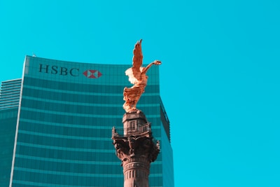 A golden angel statue stands prominently on top of a tall column against a backdrop of a modern glass skyscraper with the HSBC logo visible. The sky is clear and bright blue, providing a striking contrast to the statue and building.