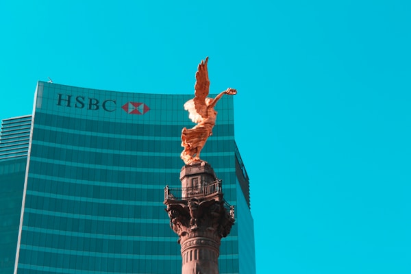 A golden angel statue stands prominently on top of a tall column against a backdrop of a modern glass skyscraper with the HSBC logo visible. The sky is clear and bright blue, providing a striking contrast to the statue and building.