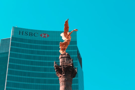 A golden angel statue stands prominently on top of a tall column against a backdrop of a modern glass skyscraper with the HSBC logo visible. The sky is clear and bright blue, providing a striking contrast to the statue and building.
