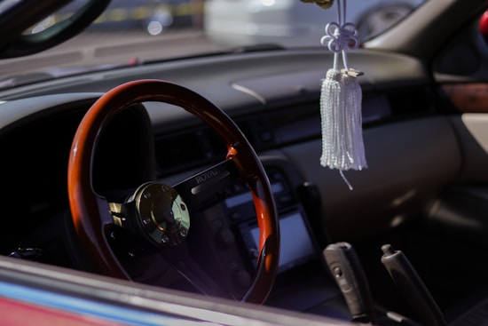 A car interior featuring a wooden steering wheel with a metallic center, adorned with a rearview hanging decoration. The dashboard and center console are visible, highlighting a modern, clean design.