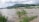 A flooded landscape with water covering a large area. Green shrubs and grass partially submerged, while a mountain range extends across the horizon under a cloudy sky. Several people are wading through the water, indicating a flood situation.