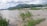 A flooded landscape with water covering a large area. Green shrubs and grass partially submerged, while a mountain range extends across the horizon under a cloudy sky. Several people are wading through the water, indicating a flood situation.
