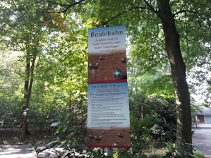 A sign for a boule court is displayed in an outdoor setting surrounded by trees and greenery. The sign contains information and rules in German, with sections labeled 'Boulebahn' and 'Spielregeln Boule'. The sky visible through the leaves appears bright, indicating daylight.