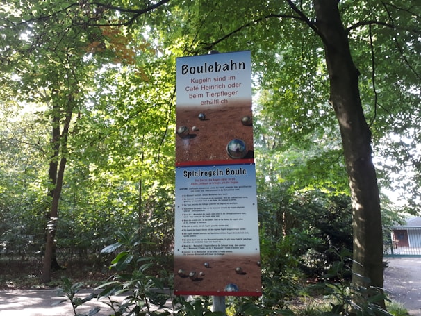 A sign for a boule court is displayed in an outdoor setting surrounded by trees and greenery. The sign contains information and rules in German, with sections labeled 'Boulebahn' and 'Spielregeln Boule'. The sky visible through the leaves appears bright, indicating daylight.