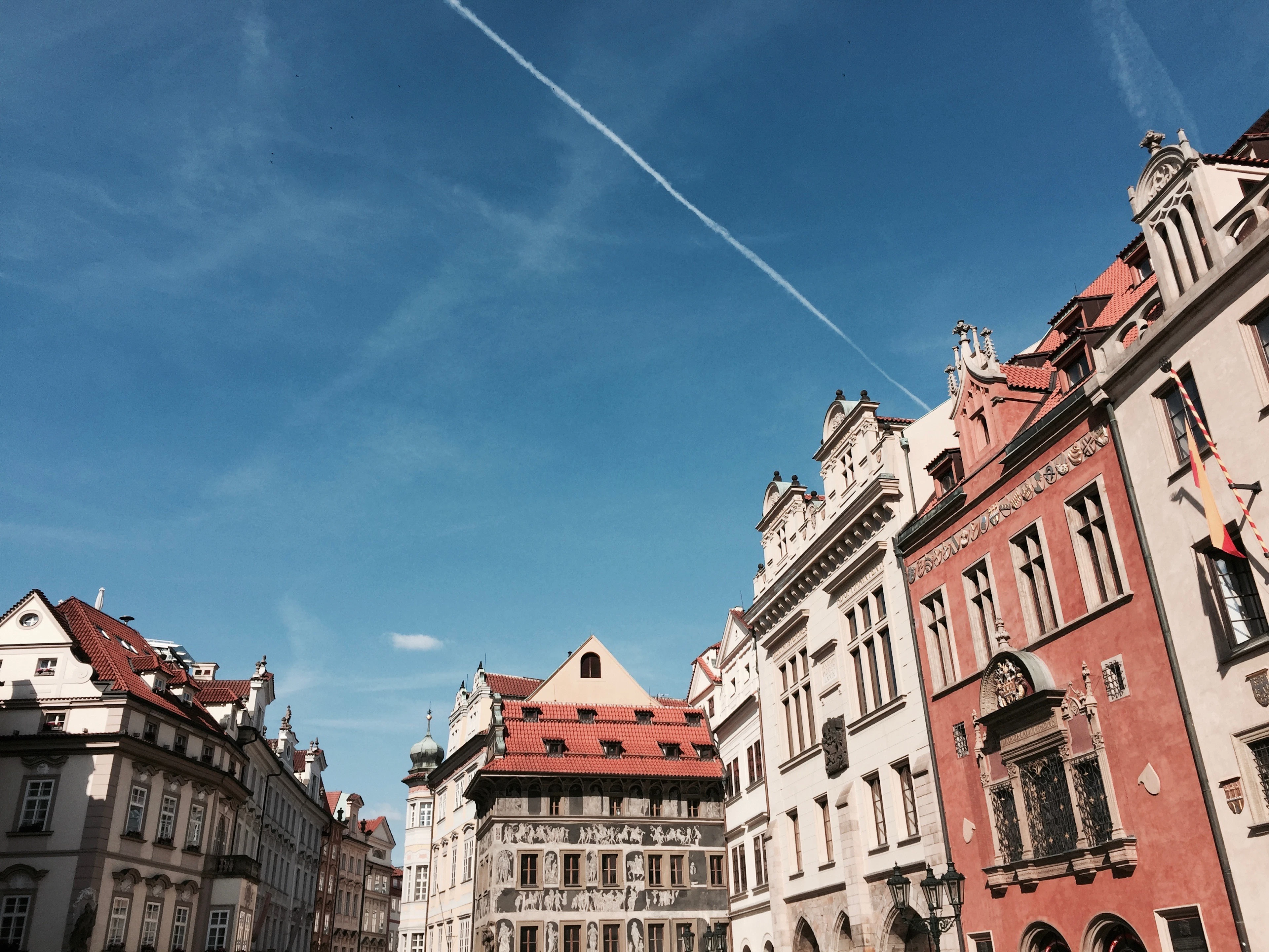 A row of Prague Old Town houses seen from below — a deep red renaissance facade, a white sgraffito-covered gable and cream baroque frontages under red-tiled roofs against a bright blue sky.