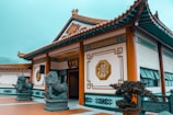 An ornate traditional Chinese building with intricate designs on the walls and roof. Two stone lion statues flank the entrance, which is open and adorned with elegant Chinese characters and geometric patterns. The surrounding area is well-maintained with plants and a tiled courtyard.