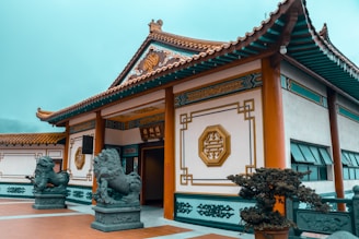 An ornate traditional Chinese building with intricate designs on the walls and roof. Two stone lion statues flank the entrance, which is open and adorned with elegant Chinese characters and geometric patterns. The surrounding area is well-maintained with plants and a tiled courtyard.