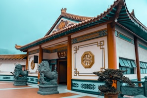 An ornate traditional Chinese building with intricate designs on the walls and roof. Two stone lion statues flank the entrance, which is open and adorned with elegant Chinese characters and geometric patterns. The surrounding area is well-maintained with plants and a tiled courtyard.