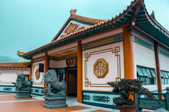 An ornate traditional Chinese building with intricate designs on the walls and roof. Two stone lion statues flank the entrance, which is open and adorned with elegant Chinese characters and geometric patterns. The surrounding area is well-maintained with plants and a tiled courtyard.