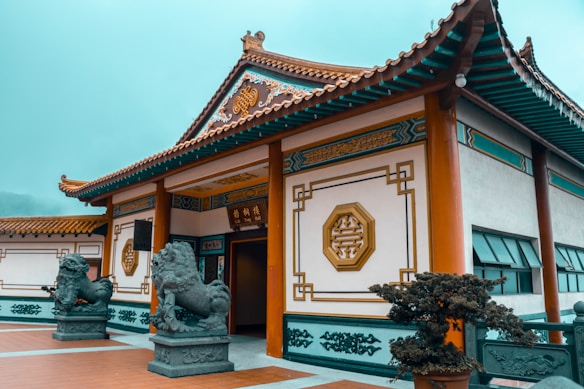An ornate traditional Chinese building with intricate designs on the walls and roof. Two stone lion statues flank the entrance, which is open and adorned with elegant Chinese characters and geometric patterns. The surrounding area is well-maintained with plants and a tiled courtyard.