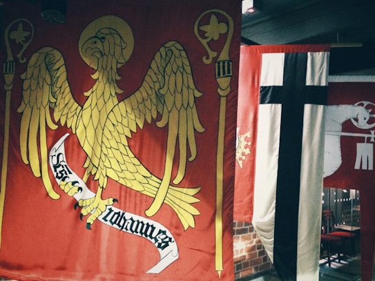 A large red banner with a golden eagle holding a scroll, surrounded by two staffs, is prominently displayed. Next to it is a vertical banner with a black cross on a white and red background. Another red flag, partially visible, features a white animal holding a staff.