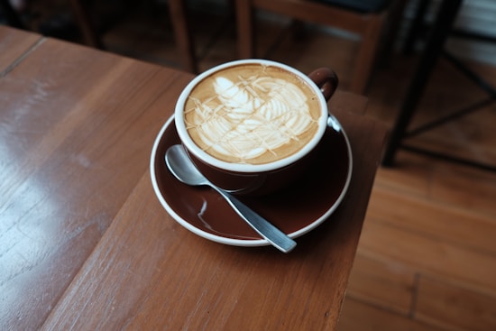 A latte in a brown cup and saucer, with a spoon, placed on a wooden table. The latte has a caramel drizzle art on top.