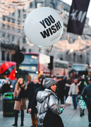 A person in a shiny silver jacket and gray beanie holds a white balloon with the words 'YOU WISH!' printed on it. They are in a busy urban setting with blurred background showing people walking, red double-decker buses, and festive lights strung across the street.