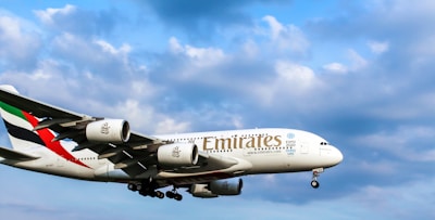 A large commercial airplane with 'Emirates' branding is flying in a clear blue sky with some fluffy clouds. The aircraft has a distinct color scheme featuring red and green on its tail. The airplane's landing gear is visible.