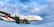 A large commercial airplane with 'Emirates' branding is flying in a clear blue sky with some fluffy clouds. The aircraft has a distinct color scheme featuring red and green on its tail. The airplane's landing gear is visible.