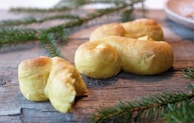 Golden-brown baked pastries with a soft texture are placed on a rustic wooden surface. Fresh pine branches are scattered around, adding a festive, natural touch.