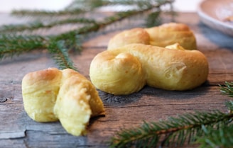 Golden-brown baked pastries with a soft texture are placed on a rustic wooden surface. Fresh pine branches are scattered around, adding a festive, natural touch.