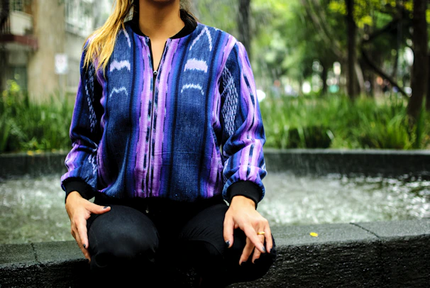 Woman wearing a purple and green gradient handmade jacket outdoors with sunlight highlighting textures.