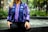 A person wearing a colorful jacket with blue and purple patterns sits by a fountain. The background features lush greenery and soft lighting, creating a serene outdoor environment. The image has a balanced focus between the detailed textile design of the jacket and the natural backdrop.