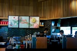 A modern coffee shop counter with a sleek design, featuring wooden slats on the ceiling and dark tiled walls. The area is equipped with coffee brewing machines, syrup dispensers, and a variety of cups and lids. There are posters with festive imagery and the phrase 'Good Together'. Two individuals appear focused on their work behind the counter.