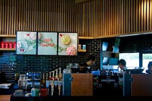 A modern coffee shop counter with a sleek design, featuring wooden slats on the ceiling and dark tiled walls. The area is equipped with coffee brewing machines, syrup dispensers, and a variety of cups and lids. There are posters with festive imagery and the phrase 'Good Together'. Two individuals appear focused on their work behind the counter.