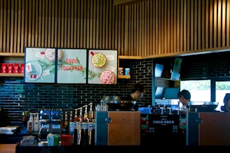 A modern coffee shop counter with a sleek design, featuring wooden slats on the ceiling and dark tiled walls. The area is equipped with coffee brewing machines, syrup dispensers, and a variety of cups and lids. There are posters with festive imagery and the phrase 'Good Together'. Two individuals appear focused on their work behind the counter.