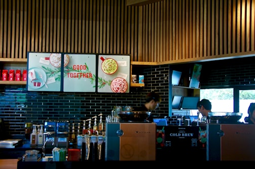 A modern coffee shop counter with a sleek design, featuring wooden slats on the ceiling and dark tiled walls. The area is equipped with coffee brewing machines, syrup dispensers, and a variety of cups and lids. There are posters with festive imagery and the phrase 'Good Together'. Two individuals appear focused on their work behind the counter.
