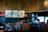 A modern coffee shop counter with a sleek design, featuring wooden slats on the ceiling and dark tiled walls. The area is equipped with coffee brewing machines, syrup dispensers, and a variety of cups and lids. There are posters with festive imagery and the phrase 'Good Together'. Two individuals appear focused on their work behind the counter.