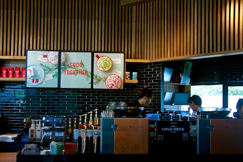 A modern coffee shop counter with a sleek design, featuring wooden slats on the ceiling and dark tiled walls. The area is equipped with coffee brewing machines, syrup dispensers, and a variety of cups and lids. There are posters with festive imagery and the phrase 'Good Together'. Two individuals appear focused on their work behind the counter.