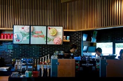 A modern coffee shop counter with a sleek design, featuring wooden slats on the ceiling and dark tiled walls. The area is equipped with coffee brewing machines, syrup dispensers, and a variety of cups and lids. There are posters with festive imagery and the phrase 'Good Together'. Two individuals appear focused on their work behind the counter.