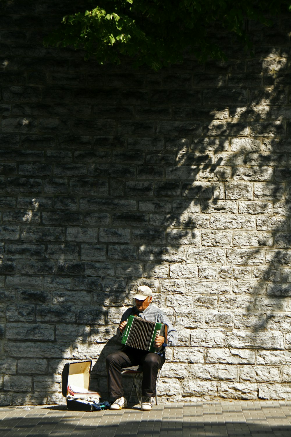 man wearing grey shirt playing accordion instrument while sitting near concrete wall and case during daytime