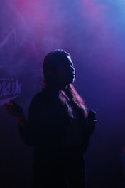 A silhouette of a person holding a microphone, with a backdrop of swirling pink and blue smoke or fog. The lighting creates a dramatic and mysterious atmosphere.