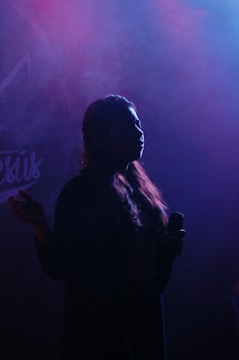 A silhouette of a person holding a microphone, with a backdrop of swirling pink and blue smoke or fog. The lighting creates a dramatic and mysterious atmosphere.